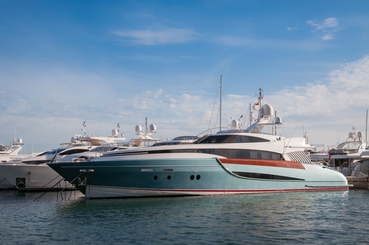 about-02 A collection of sleek yachts docked at a serene marina with a bright blue sky above.
